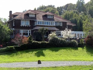 A large, two-story Craftsman-style house with a brown exterior, surrounded by green trees, a manicured garden, and a blooming white tree, overlooking a sunlit grassy lawn with a circular walking path in front.