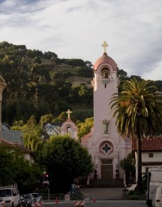 A pink stucco church with gold crosses and a bell tower stands in front of a tree-covered hillside, framed by palm trees and other buildings on a partly cloudy day.