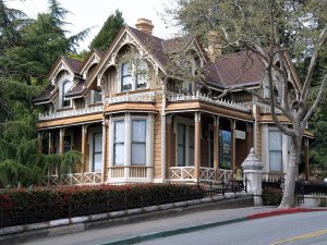 Victorian-style house with intricate woodwork, bay windows, and a wraparound porch, surrounded by trees and a black iron fence, located on a sloped street.