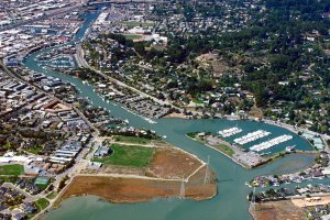 Aerial view of a coastal town with winding waterways, marinas filled with boats, green parks, neighborhoods, and a mix of residential and commercial buildings, surrounded by trees and natural wetlands.