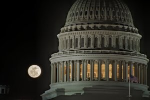 A full moon glows brightly in the night sky beside the illuminated dome of the United States Capitol Building. The architecture features elegant columns and detailed stonework, reminiscent of a grand Federal job interview setting. The American flag is visible in the lower right corner.