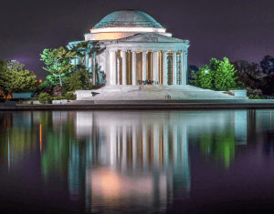 A nighttime view of the Jefferson Memorial in Washington, D.C., illuminated against the dark sky. The neoclassical structure's white columns and dome are reflected in the calm waters of the Tidal Basin, much like reflecting on a successful federal job interview with a sense of serenity. Surrounding trees are also visible.