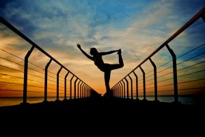 Silhouette of a person performing a yoga pose on a pier at sunrise or sunset. The person stands with one leg lifted and held by one hand while the other arm is extended. The sky, painted with shades of orange, yellow, and blue, reflects in the calm water—a serene moment before diving into careers in government.