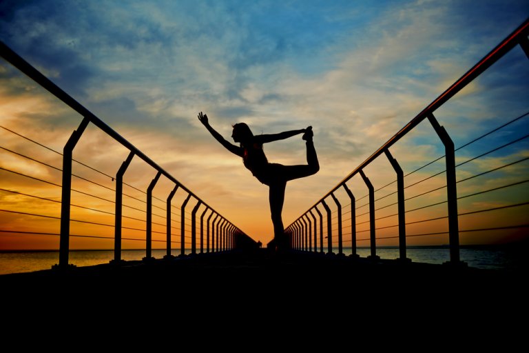 Silhouette of a person performing a yoga pose on a pier at sunrise or sunset. The person stands with one leg lifted and held by one hand while the other arm is extended. The sky, painted with shades of orange, yellow, and blue, reflects in the calm water—a serene moment before diving into careers in government.