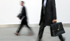 Two individuals in business attire are walking in opposite directions with blurred motion, suggesting haste or movement. Both are carrying briefcases and are set against a plain grey background. The picture captures the hectic nature of a job search within a professional environment.