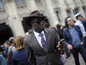 A well-dressed man wearing a brown hat, sunglasses, a brown suit, a white shirt, and a patterned tie is keeping cool while looking at his smartphone in the crowd. The background features an ornate building with a mix of men and women around him.