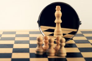 A group of white pawns on a chessboard are positioned in front of a round mirror. In the mirror's reflection, they see a white king piece, embodying the essence of leadership. The background is plain beige.