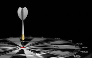 A dart is stuck precisely in the bullseye of a dartboard, showcasing sheer perfection. The dartboard is illuminated, highlighting the accuracy of the shot, while the surrounding area remains dark, drawing attention to the center of the board.