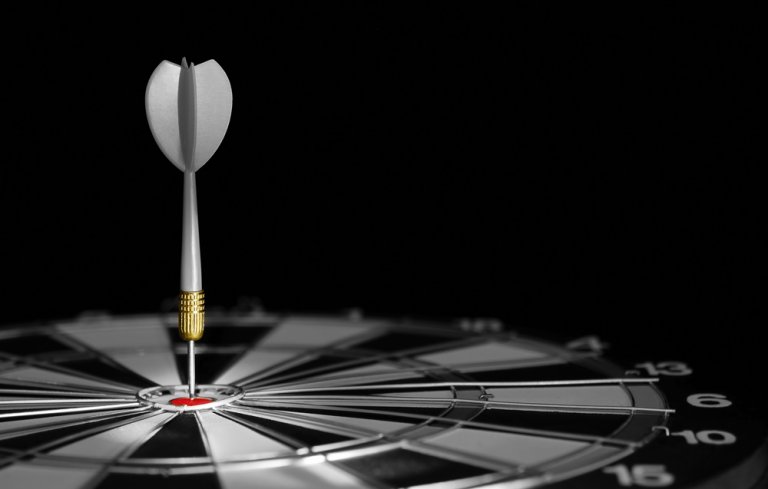 A dart is stuck precisely in the bullseye of a dartboard, showcasing sheer perfection. The dartboard is illuminated, highlighting the accuracy of the shot, while the surrounding area remains dark, drawing attention to the center of the board.