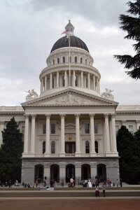 A grand, white neoclassical building stands tall with a large central dome topped by a flagpole featuring the American flag. The facade has majestic pillars and ornate sculptures. Several people are gathered near the entrance at the bottom. Trees frame the scene.