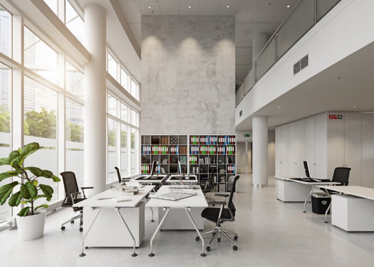 A modern, open-concept office with high ceilings and large windows allowing natural light. It features several workstations with black chairs, white desks, and computer monitors. American workers will find a large bookshelf filled with colorful binders and a potted plant.