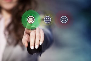 A person is pressing a green button with a smiling face icon on a touchscreen, presumably part of a survey. Two other buttons, showing a neutral face (yellow) and a sad face (red), are visible but blurred in the background, which remains out of focus.