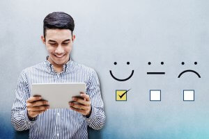 A man in a striped shirt holds a tablet and smiles while looking at it. To his right, there are four emoticons (happy, neutral, and sad). The happy emoticon is checked with a yellow box, indicating positive survey feedback or satisfaction.