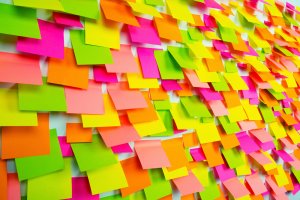 A wall is covered with a dense arrangement of vibrant sticky notes in neon pink, orange, yellow, and green. The notes overlap each other, creating a colorful and lively mosaic pattern that showcases an array of ongoing tasks.