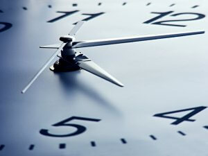 Close-up of an analog clock face showing the hands and numbers in sharp focus. The minute, hour, and second hands are prominent, casting subtle shadows on the clock face, giving it a somewhat three-dimensional look. The bold, clearly legible numbers remind one of scheduled tasks and time management.