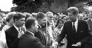 A man in a suit and tie is shaking hands with a person in the crowd, appearing as the figure of importance. Center of attention, he looks like someone you'd want to feature on your resume. Smiling faces and curious onlookers surround them against an outdoor, green background.