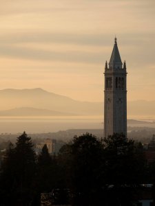 A tall clock tower stands above trees and buildings at sunset, with distant mountains and a hazy sky in the background.