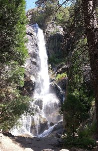 A tall, narrow waterfall cascades down a rocky cliff surrounded by lush green trees and vegetation under a clear blue sky.