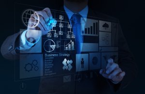 A project manager in a suit interacts with a transparent digital interface displaying various business-related icons and graphs. The interface includes charts, strategy diagrams, and key terms such as "Business Strategy" and "Innovation." The background is dark.