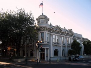 A historic two-story building with classical architectural details and columns, flying an American flag on its rooftop. Trees and streetlights frame the scene, and a car is parked nearby on a sunny day.