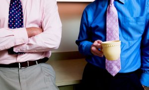 Two individuals are seen from the shoulders down, wearing dress shirts and ties—one in a pink shirt with a blue polka dot tie and the other in a blue shirt with a pink patterned tie. Exhibiting relaxed body language, the person in blue holds a beige mug as both lean against a countertop.