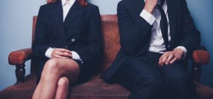 Two individuals, a woman and a man, sit side by side on a brown leather sofa against a plain background. Both are dressed in formal business attire; the woman wears a black blazer and skirt, while the man wears a black suit and tie, with his hand covering his mouth, subtly revealing their body language tension.