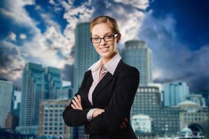 A woman wearing glasses and a black business suit stands with her arms crossed, smiling confidently. The background features a cityscape with tall buildings under a cloudy sky, highlighting her as a true leader.