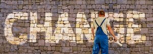 A person in blue overalls and a beige shirt is painting the word "CHANGE" on a stone wall using a roller brush. The word stands out in white against the rustic, textured beige stones. The millennial artist is facing the wall, with their back to the viewer.