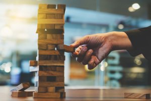 A close-up of a hand carefully removing a block from a wooden Jenga tower on a table. The background is slightly blurred, suggesting an indoor setting, likely an office or a living room. The tower appears precariously balanced, embodying the delicate balance of employee morale in workplace dynamics.