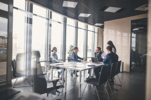 A group of five people in business attire are having a meeting in a modern office with large windows and city views. They are seated around a conference table with laptops and documents in front of them, focusing on strategies to improve employee morale. One person appears to be presenting information.