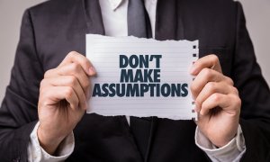 A person in a black suit and tie holds a torn piece of paper with the printed message "DON'T MAKE ASSUMPTIONS." The background is neutral, and the focus is on the hands holding the paper, as if to say, "Tell Me About Yourself" without any prior judgments.