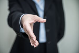 Close-up of an outstretched hand in a handshake gesture, poised to make a lasting impression. The person is wearing a dark suit with a light blue shirt underneath. The background is neutral and out of focus.