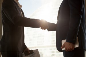 Two people in business attire shaking hands in a well-lit room with a large window in the background create a lasting impression. One person is holding documents and the other is holding a tablet, indicating a formal agreement or partnership.