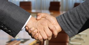Close-up of two people in business attire shaking hands, symbolizing an agreement or partnership. The background features a blurry conference room with a wooden table and chairs, evoking the setting where leaders come together to shape the future.