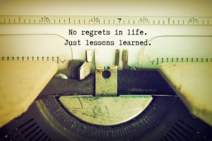 Close-up of a vintage typewriter with the text "No regrets in life. Just lessons learned." typed on the paper. The image has an aged sepia tone, evoking nostalgia and resolve. The typewriter's keys and paper holder are partially visible.