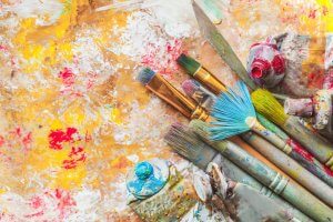 A collection of well-used paintbrushes and tubes of paint are scattered on a vibrant, abstract painted surface. The background is a mix of yellow, red, and white paint with splatters and streaks, showing an artistic resolve in this creative workspace.