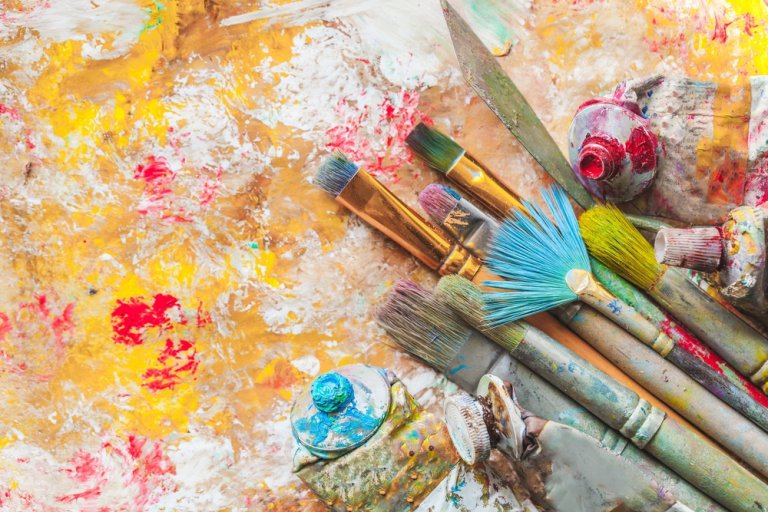 A collection of well-used paintbrushes and tubes of paint are scattered on a vibrant, abstract painted surface. The background is a mix of yellow, red, and white paint with splatters and streaks, showing an artistic resolve in this creative workspace.