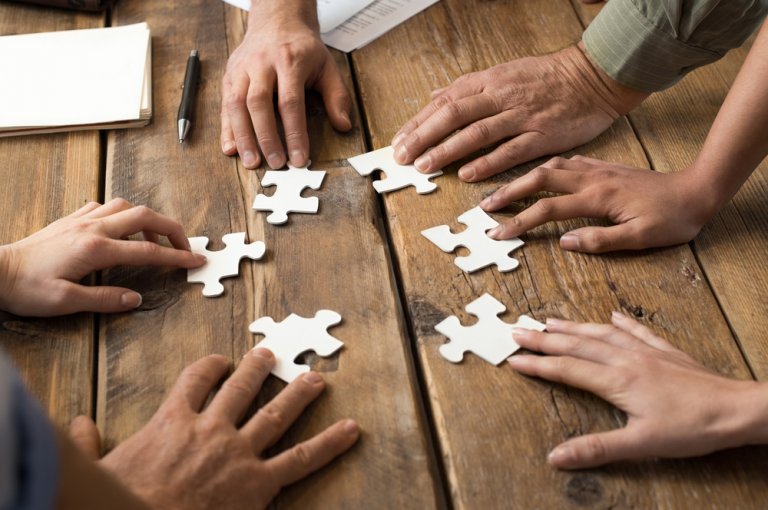 Several hands of different individuals, guided by good managers, reach out and work together to assemble white puzzle pieces on a wooden table, conveying a sense of teamwork and collaboration.