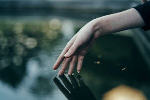 A pale hand with polished nails gently touches the surface of calm water. The background is blurred, highlighting the ripples formed by the touch. The serene and reflective nature of the scene, along with the subtle body language, suggests tranquility and connection with nature.