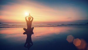 Silhouetted person sitting in a yoga pose on a serene beach at sunset, with hands raised and palms together above their head. The vibrant colors of the setting sun reflect off the calm water, creating a peaceful and meditative scene that inspires professional growth.