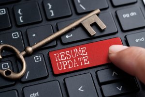 A close-up of a keyboard with a finger pressing a red key labeled "RESUME UPDATE" in white text. A large antique-style brass key is resting on the keyboard, evoking the timeless importance of updates in any resume builder's toolkit.