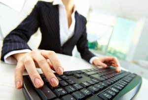 A person in a business suit is seated and typing on a black keyboard, possibly working on a resume builder. The image is taken from a low angle, focusing on the hands and keyboard. A bright office environment with large windows is visible in the background.