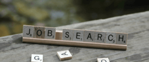 Wooden letter tiles spelling "JOB SEARCH" are arranged on a stand. Additional tiles, including "G" and "A," are scattered on a rustic wooden surface in the foreground, perhaps hinting at tools like a resume builder. The background is blurred, focusing attention on the message.
