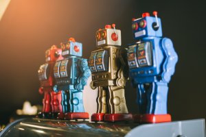 Photograph of three vintage toy robots, hinting at a robot revolution. They are lined up in a row with the first robot in red, the second in silver, and the third in blue. Each robot features colorful details and a square body design, set against a dark background with warm light from the left.