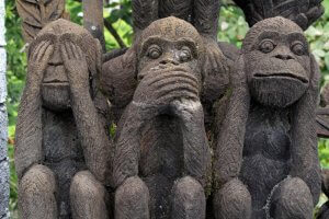 Three stone sculptures of monkeys, embodying the classic three wise monkeys motif. The first monkey covers its eyes (see no evil), the second covers its mouth (speak no evil), and the third covers its ears (hear no evil). Set against a backdrop of lush greenery, perfect for millennial mindfulness.
