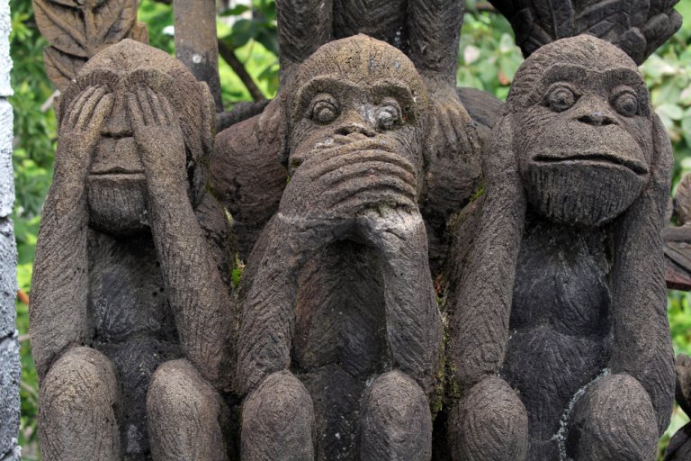Three stone sculptures of monkeys, embodying the classic three wise monkeys motif. The first monkey covers its eyes (see no evil), the second covers its mouth (speak no evil), and the third covers its ears (hear no evil). Set against a backdrop of lush greenery, perfect for millennial mindfulness.
