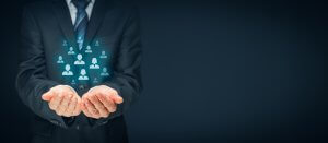 A person wearing a business suit and tie presents their open hands forward. Floating above their hands are digital icons of human figures, symbolizing concepts like team value, social network, or customer relationship management, against a dark background.