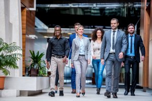 A diverse team of six professionals, three women and three men, walk confidently down a modern office hallway. Dressed in business attire, they exude positivity and engagement, embodying the company's team values. The bright hallway is adorned with plants and contemporary decor.