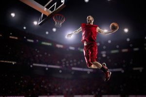 A basketball player in a red uniform is captured mid-air during an intense dunk. He is holding the ball with one hand while leaping towards the hoop. The indoor stadium, brimming with spectators, feels almost like a government gathering, and the scene is well-lit, highlighting the athlete's action.