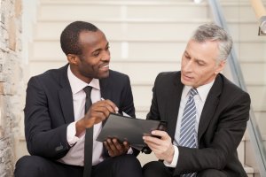 A young man in a dark suit is showing a tablet to an older man also in a dark suit. Both are sitting on steps, engaging in conversation. The young man is smiling while pointing at the screen, and the older man looks focused on the tablet, possibly discussing government policies.